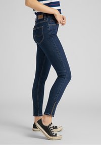 Jeans skinny azul oscuro de denim, con una cintura alta, dos bolsillos traseros y detalles con cremallera en el tobillo. Combinados con zapatillas negras.