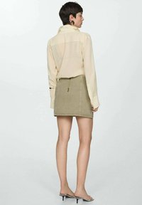 Crème lange mouwen blouse gecombineerd met een kaki mini rok. De blouse heeft een doorschijnende achterkant en strakke manchetten; de rok heeft een ritssluiting aan de voorkant.
