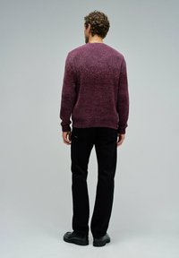 Mann mit lockigem Haar, der einen lila Pullover, schwarze Hosen und schwarze Schuhe trägt, steht mit dem Rücken zu einem einfarbigen grauen Hintergrund.