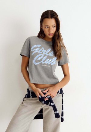 Junge Frau mit langen braunen Haaren trägt ein graues "Girls Club" T-Shirt, beige Hose und bindet einen gestreiften marineblauen Pullover um die Taille, steht vor einer weißen Wand.