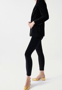 Černý sametový blejzr s úzkým střihem, spárovaný s černými elastickými kalhotami a zlatými lodičkami na podpatku. Hladká textura a elegantní design.