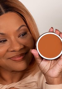 Rund kompakt av matt bronzer i en varm orange-brun nyans, innesluten i en silvermetallbehållare. Slät textur synlig på ytan.