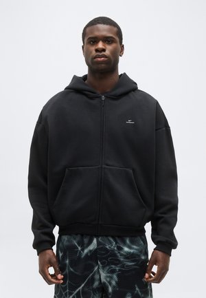 Mladý muž oblečený v čiernej mikine na zips s malým bielym logom a vzorovanými tmavými športovými šortkami, stojaci na neutrálnom pozadí.