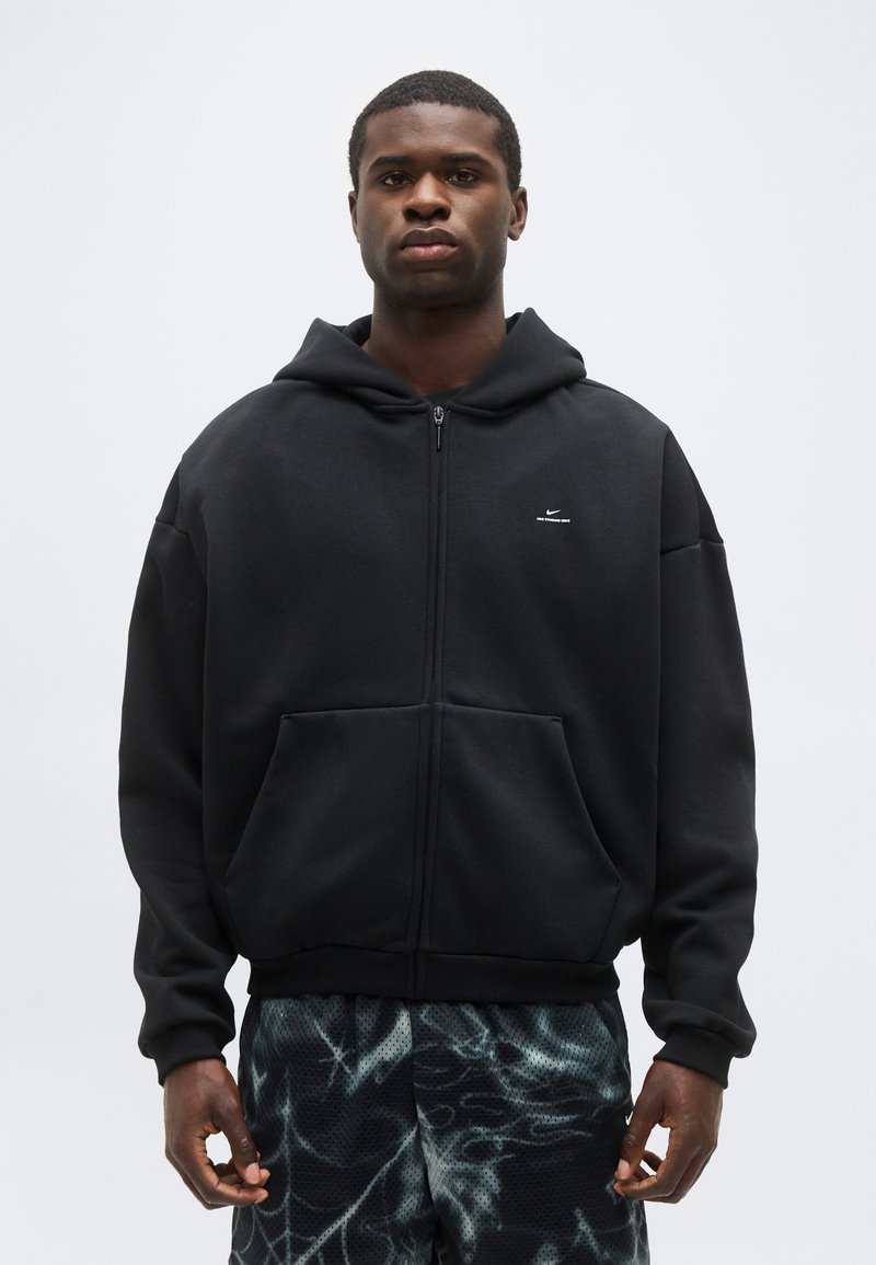 Νεαρός άνδρας που φοράει μαύρο φούτερ με φερμουάρ και μικρό λευκό λογότυπο, καθώς και σκούρα αθλητικά σορτς με σχέδια, στέκεται μπροστά σε ουδέτερο φόντο.