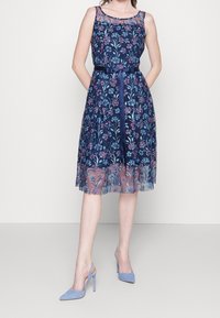 Robe florale bleu marine avec superposition transparente, brodée de fleurs roses et bleues, cintrée à la taille avec une ceinture en satin, associée à des talons bleus.