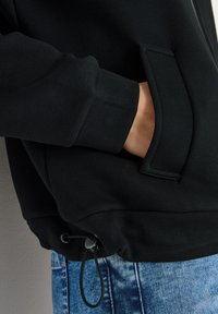 Mano en el bolsillo de un sudadera negra con cordón ajustable en el dobladillo, encima de unos jeans de mezclilla azules, contra un fondo gris.