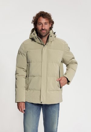 Man met krullend haar draagt een beige donzen jas met capuchon en zakken, staand met één hand in de zak tegen een effen lichte achtergrond.