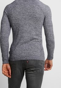 Grauer Strickpullover mit langen Ärmeln, strukturiertem Gewebe, kontrastierenden Sprengseln und figurbetontem Design. Besitzt ein kleines Logoschild nahe dem Saum.