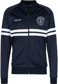 Veste de survêtement bleu marine en polyester lisse avec des rayures blanches sur les manches. Présente un zip à l'avant, un col montant et un logo brodé sur la poitrine.