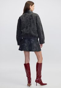 Zwarte denim jas met florale reliëf en geribbelde zoom, gecombineerd met een uitlopende donkere rok. Glanzende rode kniehoge laarzen.