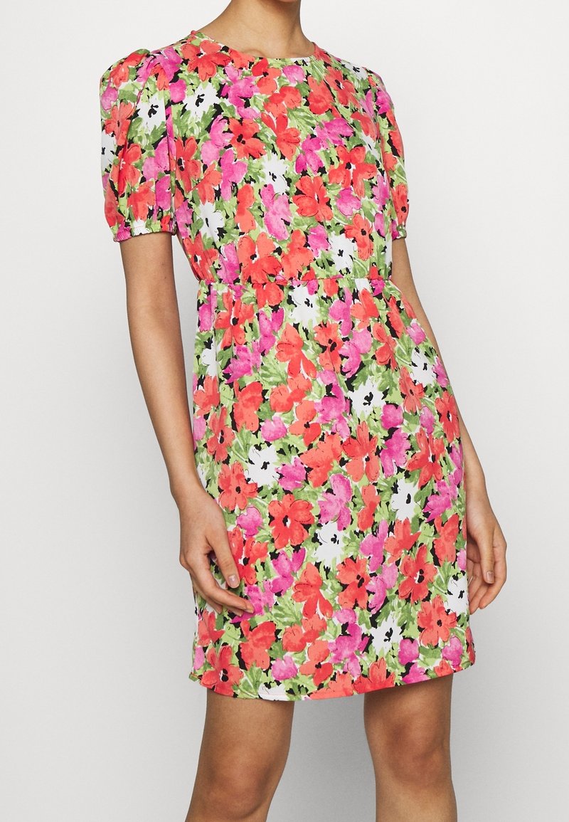 Femme portant une robe mi-longue avec un motif floral rouge et rose, des manches bouffantes et une taille cintrée sur un fond blanc uni.