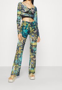 Mehrfarbiger transparenter Crop-Top mit langen Ärmeln und Bindebandverschluss, kombiniert mit einer passenden, weit geschnittenen Hose mit abstraktem Muster und Plateausandalen.