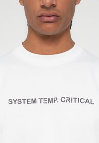 T-shirt de algodão branco com texto preto "TEMPO DO SISTEMA CRÍTICO" em uma fonte desgastada, gola redonda e mangas curtas.