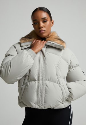 Femme ajustant le col d’une doudoune gris clair avec une bordure en fourrure beige, portant un pantalon noir sur un fond clair uni.