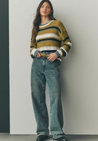 Strickpullover mit gelben, marineblauen, weißen und grauen Streifen, kombiniert mit weiten, hellblauen Jeans und schwarzen Schuhen. Strukturierter Stoff.