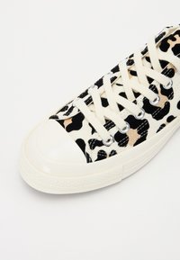 Zapatillas de lona con un diseño de estampado de leopardo en negro y beige, puntera de goma blanca y cordones beige enhebrados a través de ojales plateados.
