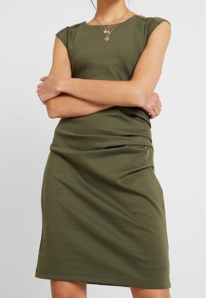 Mujer con vestido de manga corta verde oliva y collares dorados en capas, con los brazos cruzados sobre el pecho frente a un fondo liso.