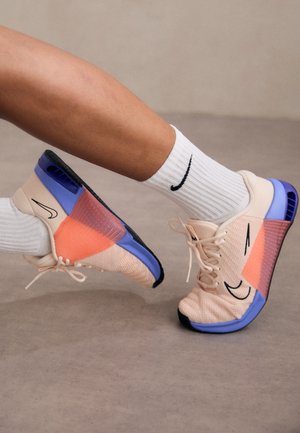 Nohy s bílými ponožkami Nike a béžovými, fialovými a oranžovými teniskami Nike na šedé podlaze, jedna noha zvednutá a ohnutá.