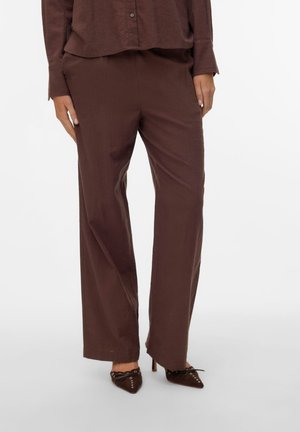 Kvinde iført løstsiddende brune bukser, matchende skjorte med knapper og spidse brune hæle med snøre- og nittedetaljer, stående mod en hvid baggrund.