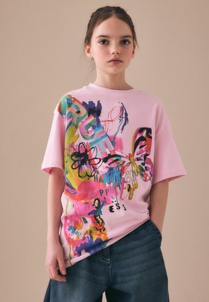 Ung person iført en løs lyserød T-shirt med farverige abstrakte blomster- og sommerfugledesign, kombineret med mørkeblå jeans.