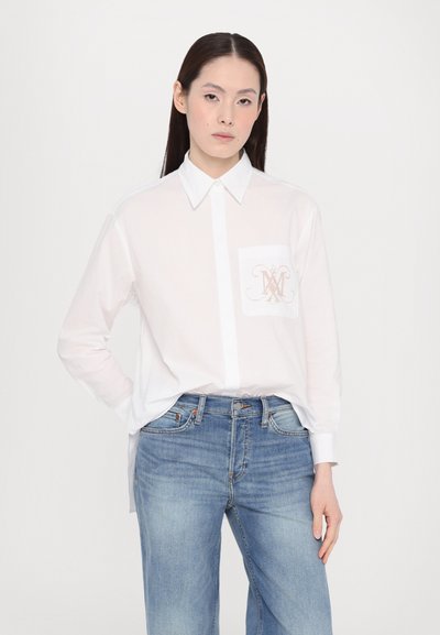 Camisa blanca de manga larga con cuello abotonado, que presenta un bolsillo en el pecho con un logotipo bordado. Combinada con unos jeans de color azul claro.