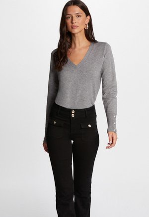 Grijze v-halstrui met ribgebreide manchetten en knoopaccenten, gecombineerd met highwaisted zwarte jeans met zijzakken en decoratieve knopen.
