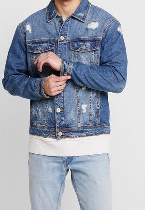 Homme ajustant le poignet d'une veste en jean bleu délavé portée sur une chemise blanche avec un jean bleu clair sur un fond uni.