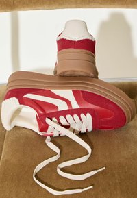 Raudoni ir kreminės spalvos sportbačiai su tekstūruotu viršumi, apvalia nosimi ir storu rudo gumo padu. Turi baltas raištelius ir sluoksniuotus dizaino akcentus.