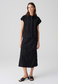 Outfit nero composto da una maglietta corta a maniche corte con texture e una gonna lunga abbinata. Il materiale sembra leggero con un motivo ondulato; accompagnato da sandali neri.