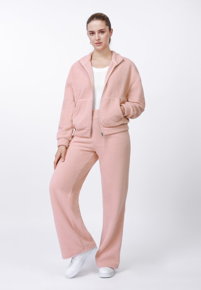 Jachetă roz din fleece cu fermoar și două buzunare frontale, asortată cu pantaloni din fleece de aceeași nuanță. Ambele piese au o textură moale și un croi lejer.