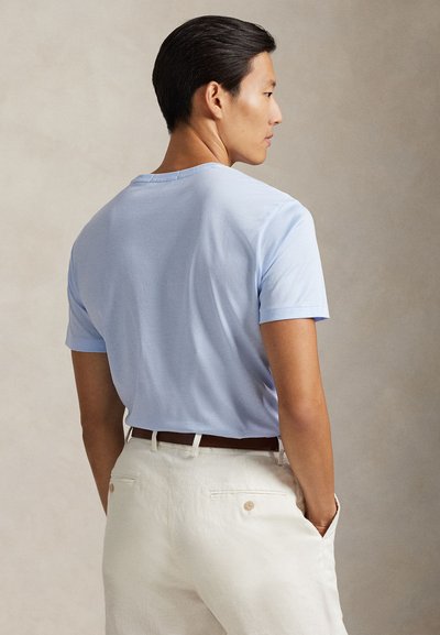 Άνδρας με σκουρόχρωμα μαλλιά, ντυμένος με ανοιχτού μπλε κοντομάνικο πουκάμισο που είναι περασμένο μέσα σε λευκό παντελόνι με καφέ ζώνη, στέκεται με τα χέρια στις τσέπες.