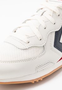 Sneaker bianco in pelle con pannelli in rete, dettagli grigi e rifiniture rosse. Presenta lacci piatti e una suola in gomma con piccoli modelli di grip.