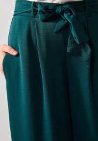 Teal, texturerade tygbyxor med knytmidja, med djupa veck och sidofickor, med fokus på funktionalitet och en svepande silhuett.