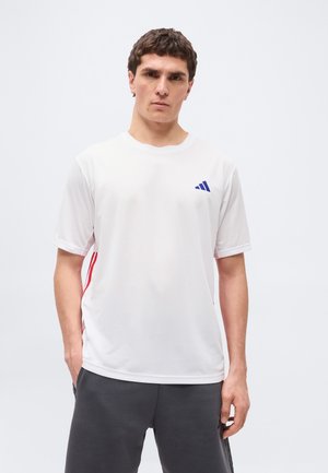 Muž v bielom športovom tričku s krátkym rukávom a malým modrým logom, v tmavosivých nohaviciach, stojaci pred jednofarebným svetlým pozadím.