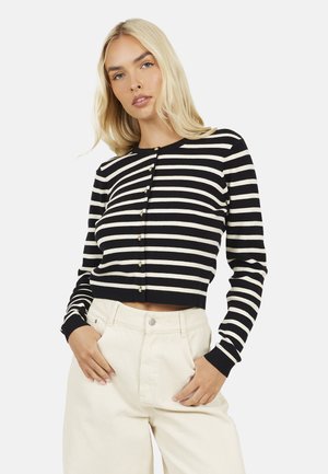 Vrouw met lang blond haar, gekleed in een zwart-wit gestreepte vest met knopen en hooggetailleerde crèmekleurige broek, staat met haar handen in haar zakken.