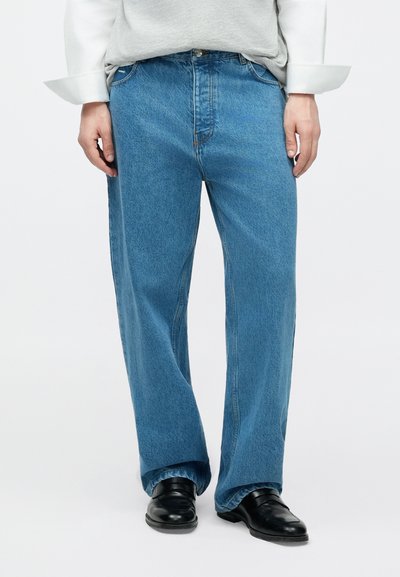 Hele sinised, sirge säärega teksapüksid klassikalise viie taskuga kujundusega. Kangal on tekstuuritud viimistlus ja kerge pleekimine. Kandetud mustade kingadega.