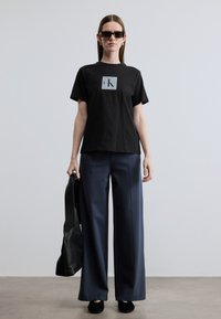 Μαύρο βαμβακερό t-shirt με γκρι λογότυπο σε τετράγωνο, συνδυασμένο με φαρδιά μπλε παντελόνια και μαύρα παπούτσια, κρατώντας μια μαύρη τσάντα ώμου.