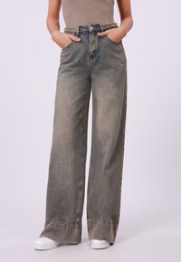 Wide-leg denim jeans in faded blue with a braided fabric belt, two front pockets, and a slightly distressed hem. White sneakers below.
