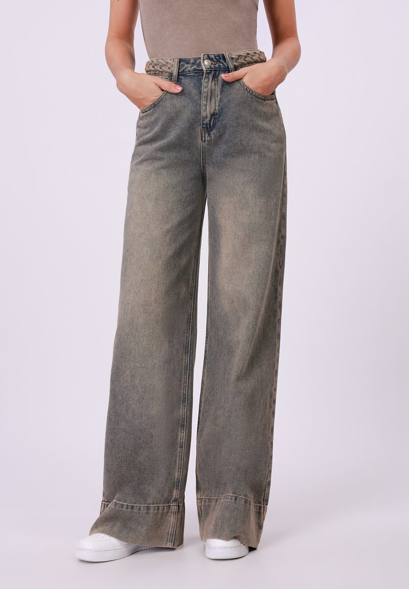 Wide-leg denim jeans in faded blue with a braided fabric belt, two front pockets, and a slightly distressed hem. White sneakers below.