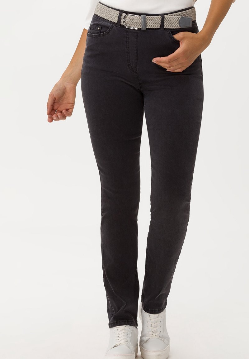 Femme portant un jean slim noir avec une ceinture beige tressée et des baskets blanches, mains près des poches.