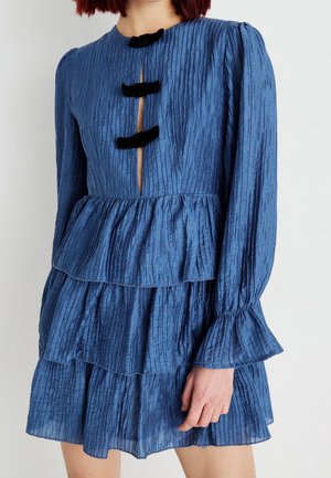 Donna che indossa un vestito blu testurizzato a maniche lunghe con balze a più livelli e chiusure a fiocco in velluto nero sul petto.