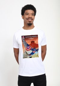 Bílý tričko s barevným potiskem z komiksu Captain America, zobrazující postavu v modré a červené s štítem a létající, na béžovém pozadí.