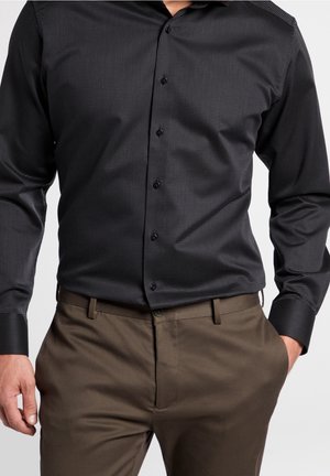 Uomo che indossa una camicia aderente con bottoni e motivo scuro infilata in pantaloni eleganti marroni, con una mano in tasca.