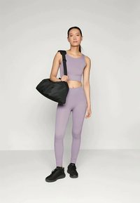 Μωβ αθλητικό crop top και κολάν σε συνδυασμό με μαύρα αθλητικά παπούτσια. Μια μαύρη τσάντα duffel με λουρί ώμου κρατιέται με το ένα χέρι.