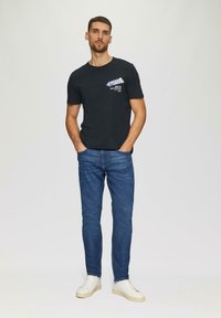 Camiseta de algodón negra con texto blanco, jeans de mezclilla azul y zapatillas beige, de pie contra un fondo claro.