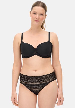 Mujer con sujetador negro con aros y bragas de encaje de talle alto, de pie contra un fondo liso claro, mirando al frente.