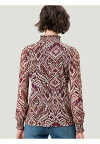 Blouse à manches longues avec un col à volants, arborant un tissu à motifs multicolores dans des teintes de bordeaux, crème et tons terreux.