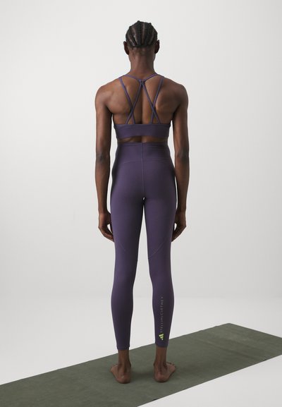 Sutiã desportivo roxo com costas em tiras, combinado com leggings de cintura alta apresentando um detalhe em logotipo na perna esquerda. Textura suave.