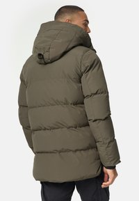 Veste matelassée verte olive avec capuche, présentant un design matelassé, des poignets élastiques et un détail de sangle au dos. Texture lisse, coupe mi-longue.