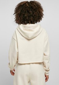 Hoodie court crème avec capuche, design oversized, poignets côtelés et texture douce. Porté avec un pantalon assorti ; la vue de dos montre les détails de l'ourlet.